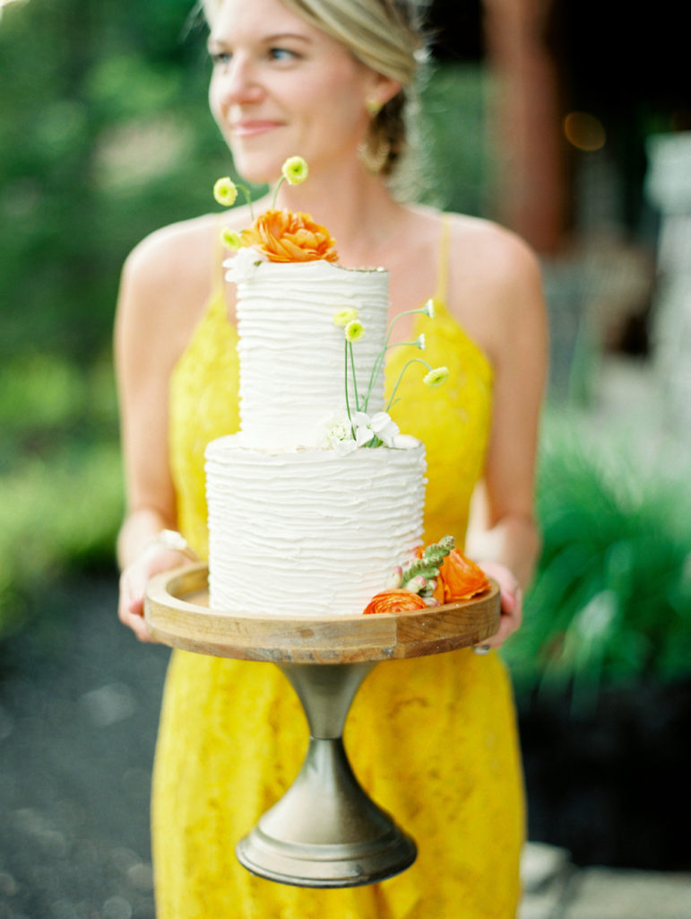 woman in yellow dress holding wedding cake with flowers by Enuf Chocolates | Mary Dougherty