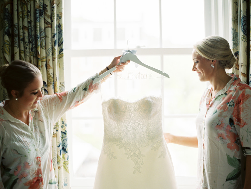 bridesmaids holding wedding dress in front of window at Otesaga | Mary Dougherty