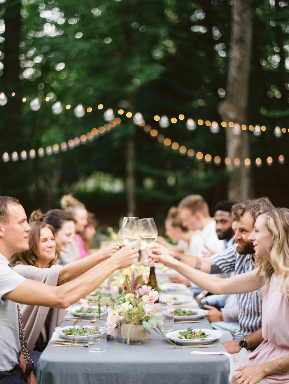 guests cheers Living roots wine at a long table wedding rehearsal dinner outside under string cafe lights | Mary Dougherty Workshop Saranac Lake New York