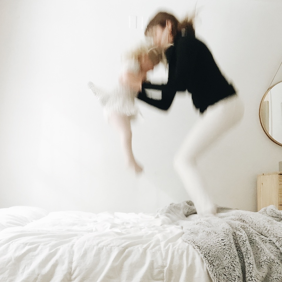 mom holds baby daughter and jumps on bed in minimalist bedroom motion blur on iphone photography by Mary Dougherty