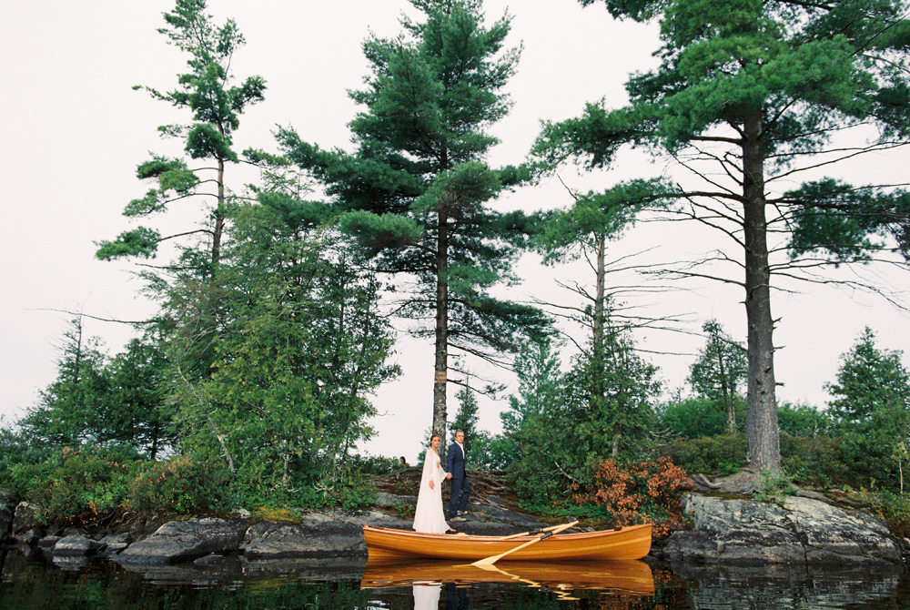 bride and groom stand on island with wooden boat after paddling out for elopement in Adirondacks in saranac Lake by Mary Dougherty