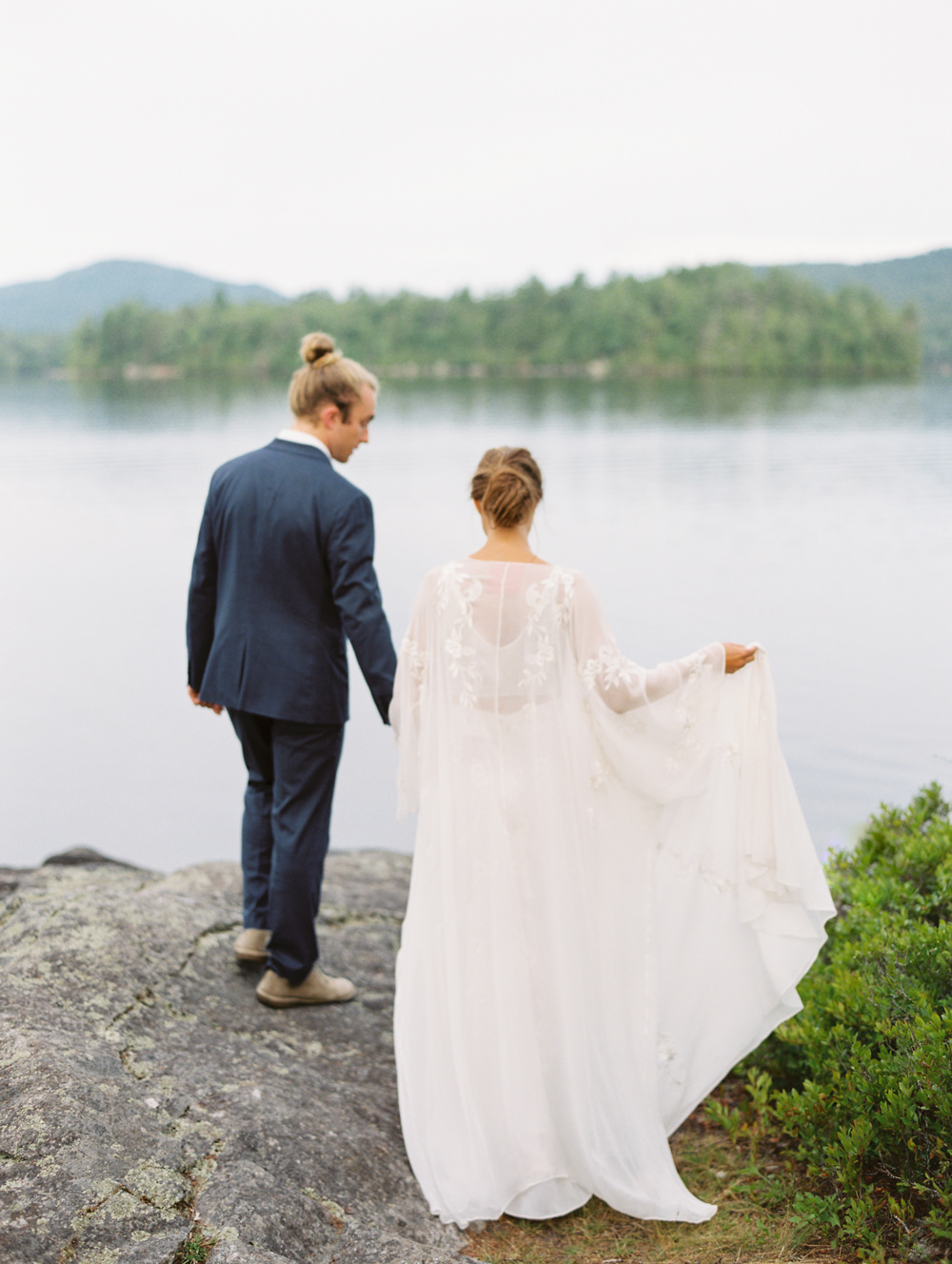 bride and groom walk on rocks towards water with long cape wedding dress by L'eto Bridal by Mary Dougherty