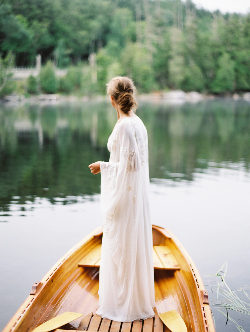 bride with perfect messy bun and long cape wedding dress stands in wooden boat looking towards lake in Adirondack Island Elopement by Mary Dougherty