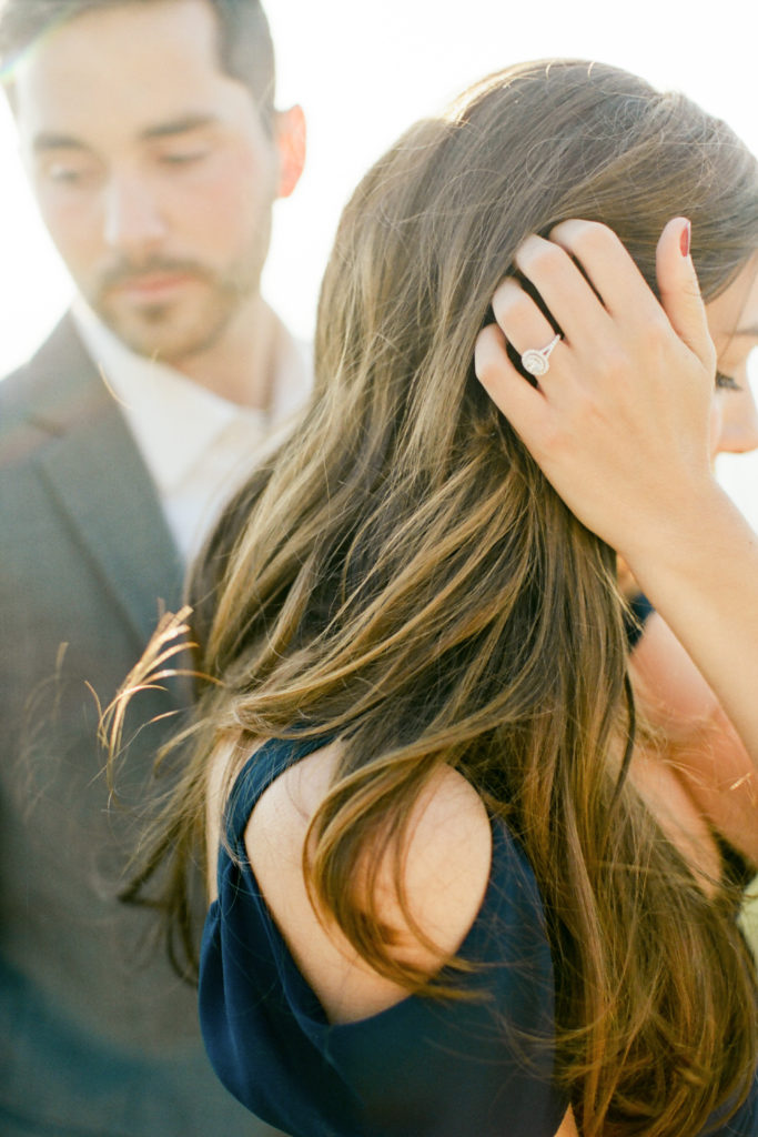 engagement ring on hand of woman brushing hair back with fiance standing behind looking aside | Whiteface mountain engagement by Mary Dougherty