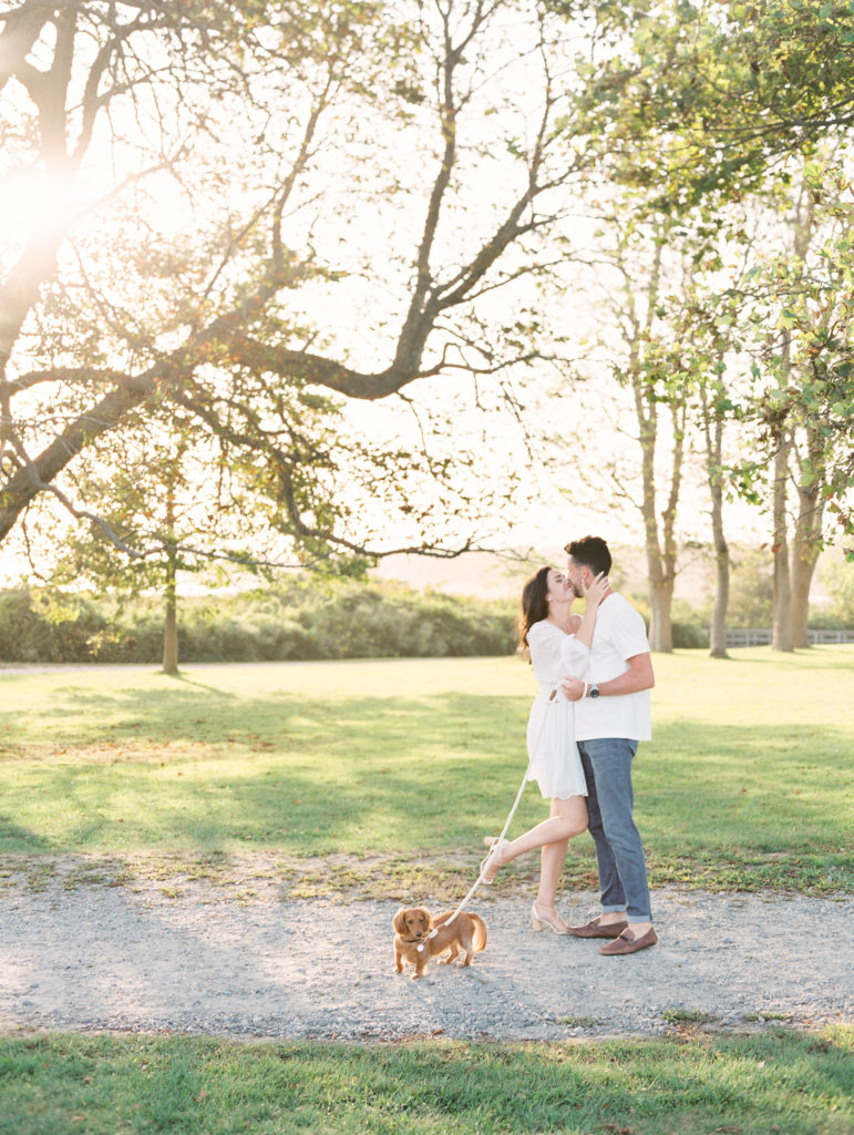 Bride and groom-to-be embracing in a park with their Dachshund for an engagement photo shoot