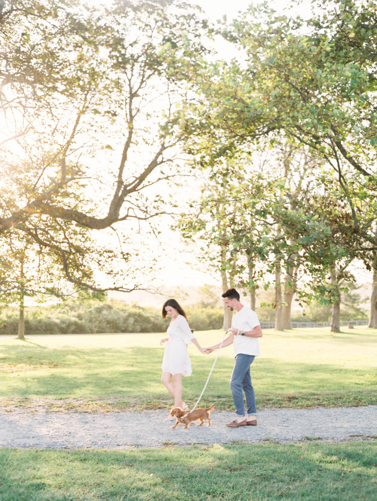 Engagement photo of a couple holding hands while walking their dog through the park