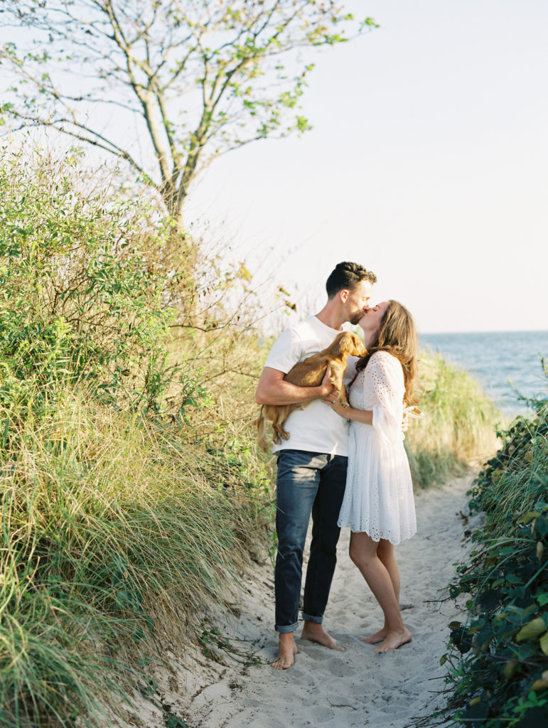 Couple embracing in the sand while holding their Dachshund in a casual beach engagement photo