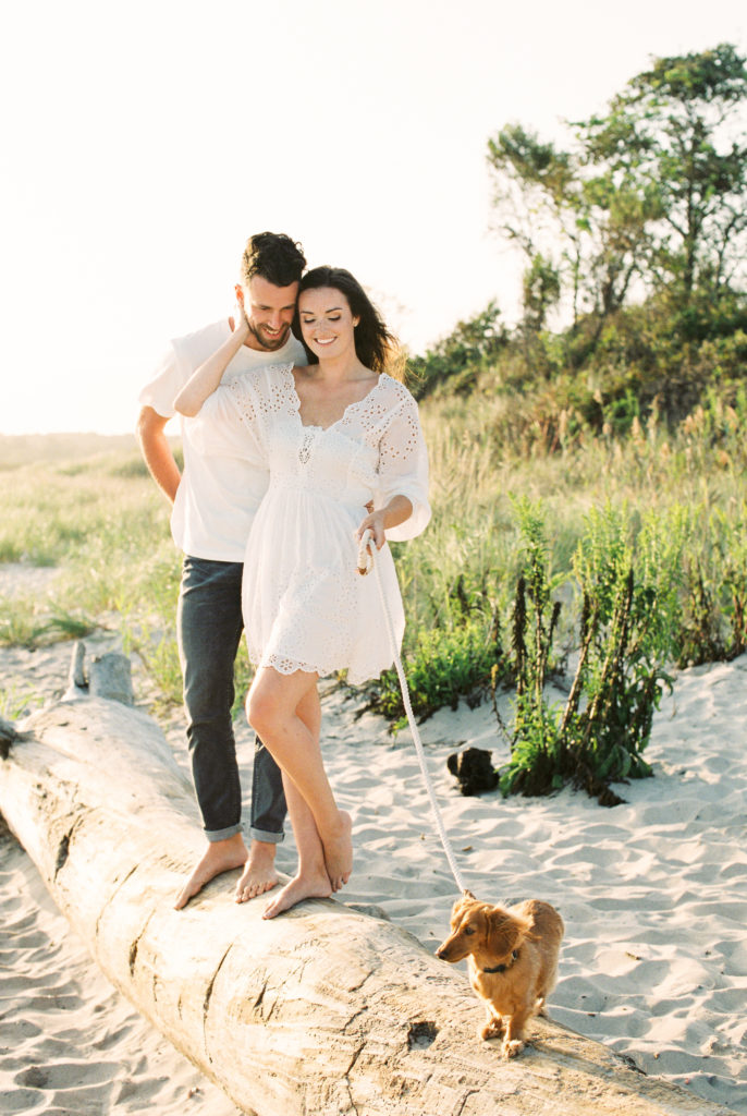 Couple standing with their dog on a fallen tree in the sand posing for engagement photos at the beach