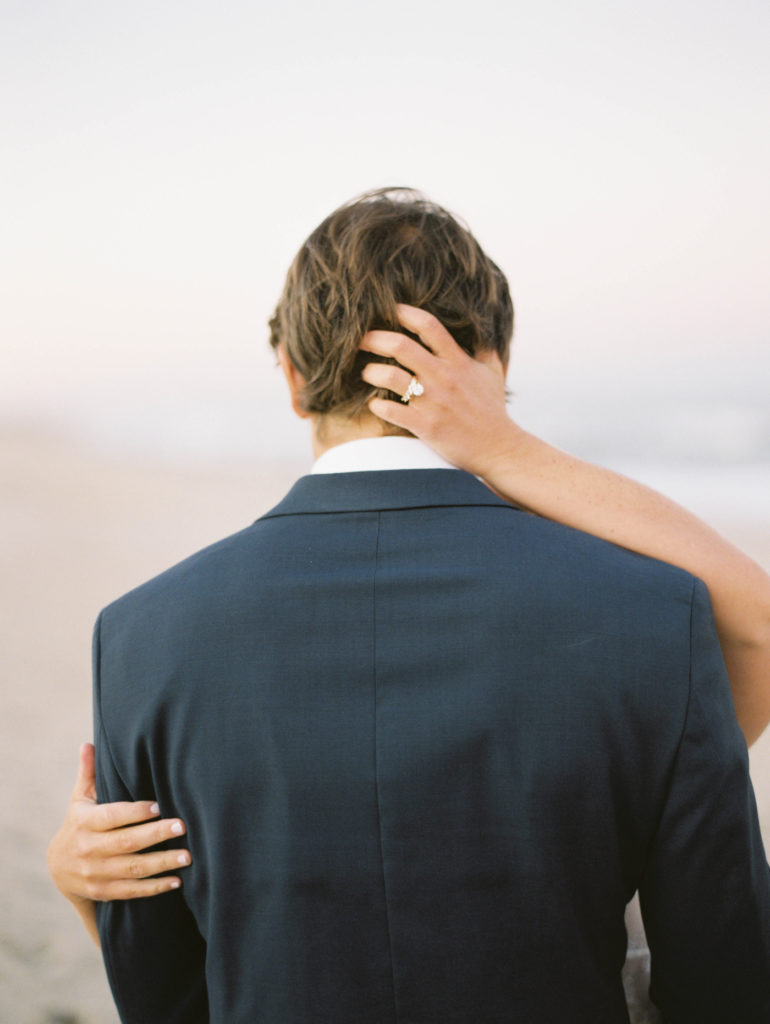 Bride has hands in grooms hair facing away from camera at the beach | editorial wedding photograph by film photographer Mary Dougherty