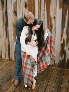 Couple embraces with plaid blanket while standing outside of wooden cabin at Gather Greene image by Mary Dougherty