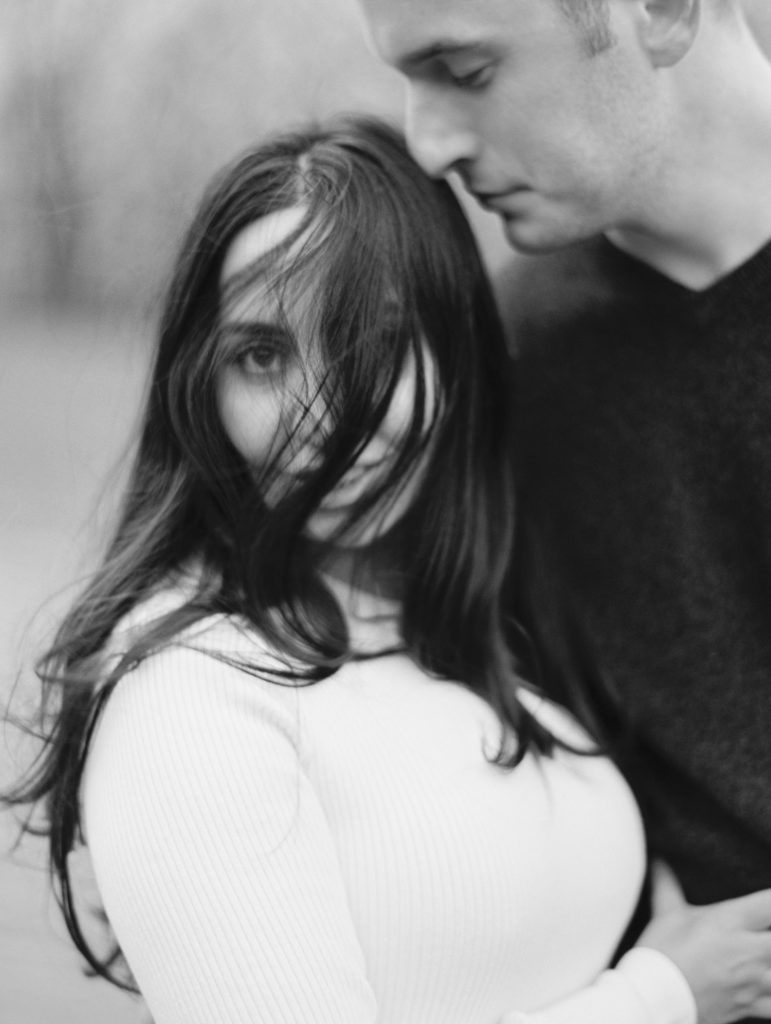 Black and white photo of man embracing woman with tousled hair in outdoor engagement photos 