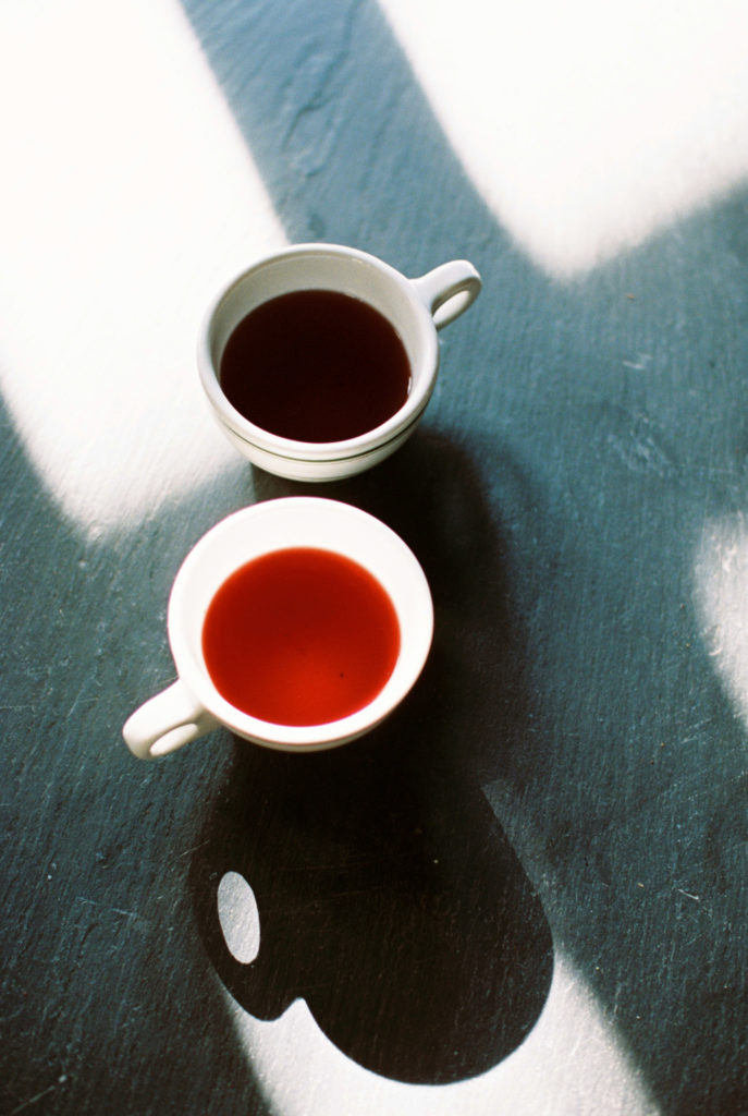 two cups of tea one red and one dark red sit on slate table with window light streaming over