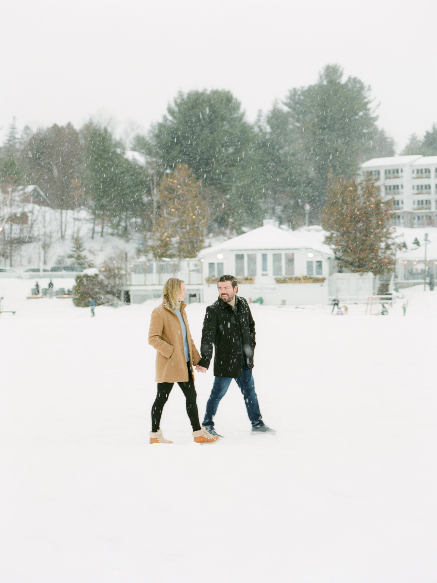 A couple walks holding hands across frozen Mirror Lake in Lake Placid while it snows. Photograph by Mary Dougherty