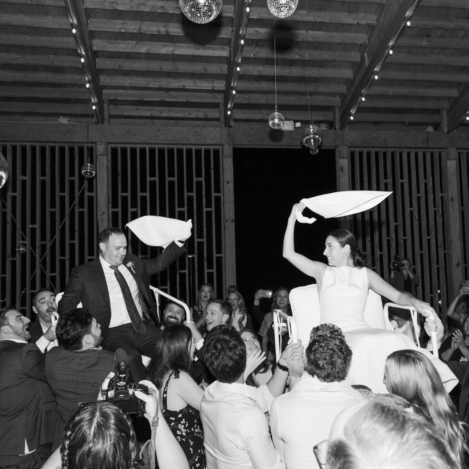 bride and groom dance the hora and are lifted in the air on chairs with napkins waving around their heads as they look at each other.