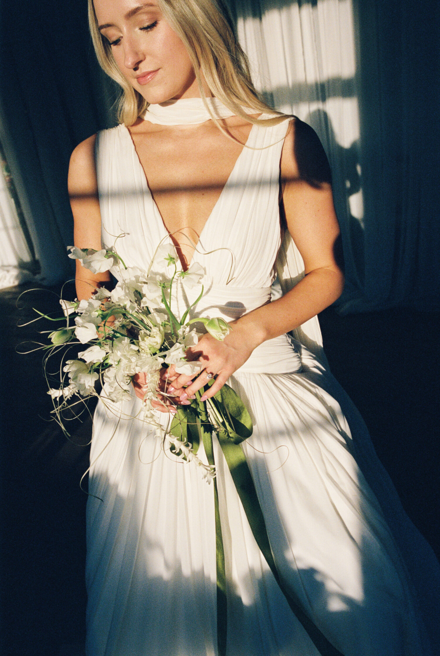 film photograph of a bride in window light with direct sun shining on bouquet of flowers that is overgrown and a little wild