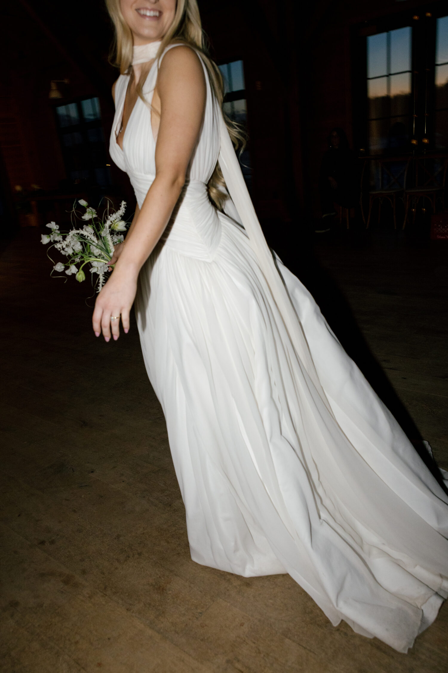photo of a bride smiling and walking inside a building with a glimpse of sunset and flash photography showing her dress and bouquet